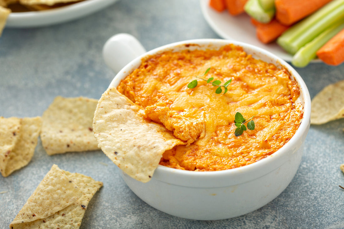 Baked buffalo chicken dip bubbling in a white dish with melted cheddar, served with tortilla chips, celery and carrot sticks
