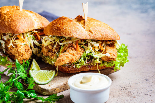 Crispy fried chicken sandwiches piled with coleslaw on toasted buns, served on a wooden board with a cup of dipping sauce