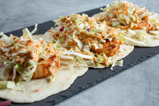 Three sweet and spicy shrimp tacos on soft tortillas topped with crunchy cabbage slaw and a peach crema drizzle, served on a black slate board